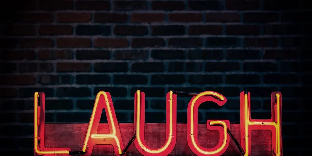 Leuchtschrift LAUGH in Rot und Gelb vor einer dunklen Backsteinwand