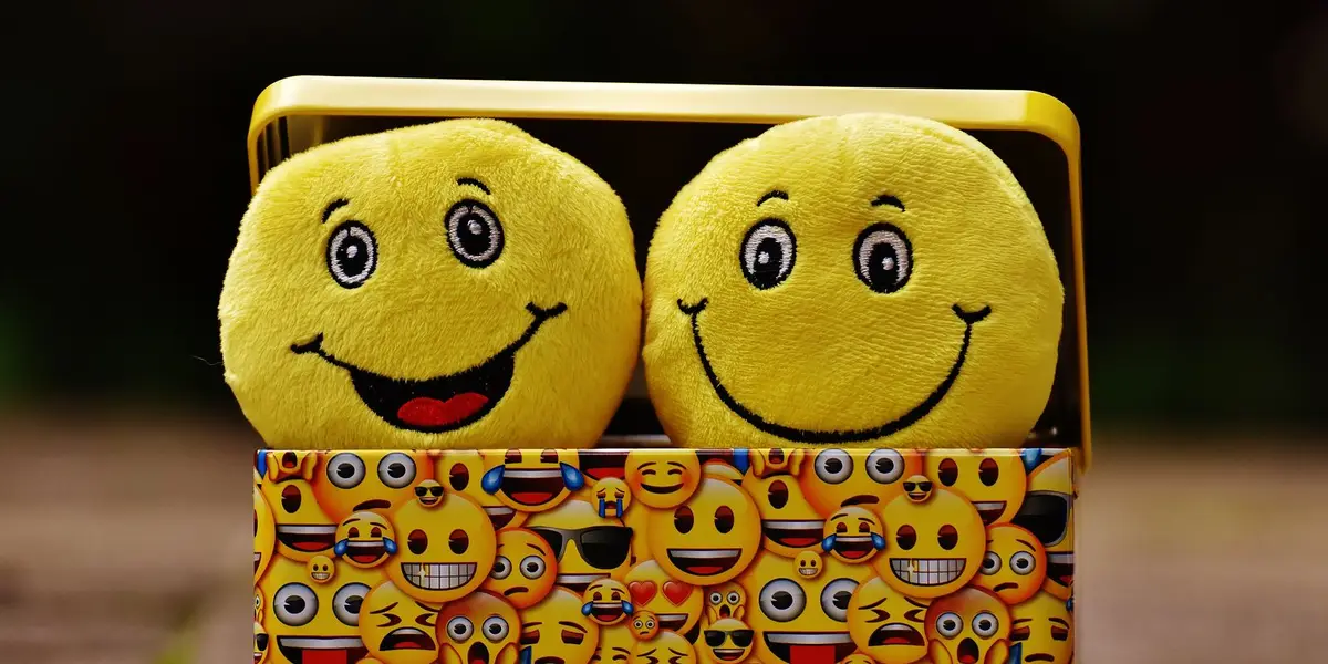 Zwei lachende Plüsch-Smileys schauen aus einer gelben Dose mit Emoji-Muster